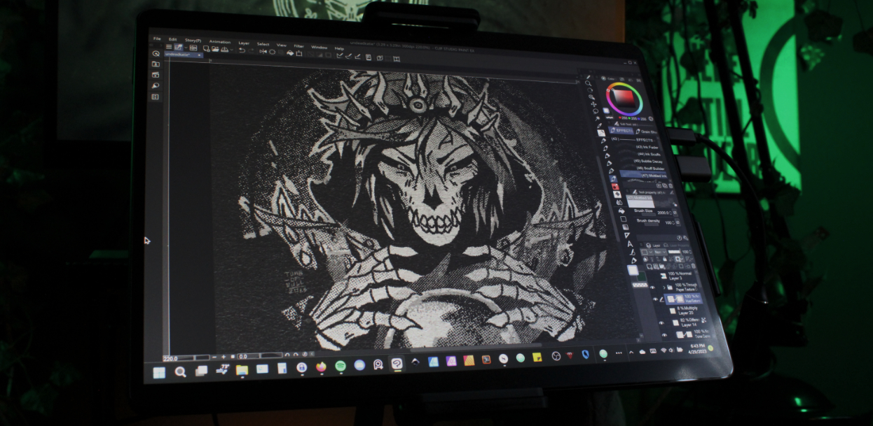 Photo of a desk with digital drawing tools set up, including a computer with extra screen. On the screen is a digital illustration of a lich queen in progress.