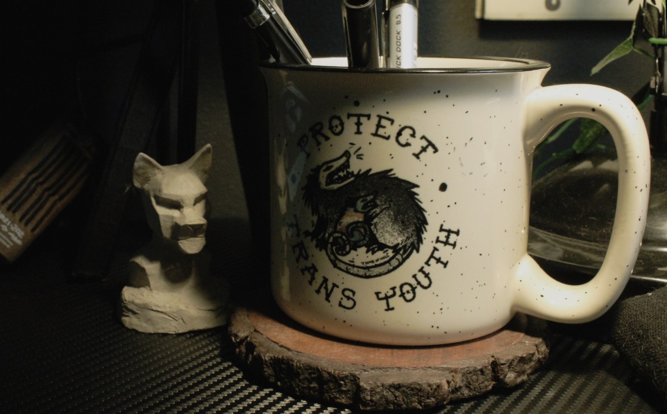 Photo of a white mug on a wood coaster. The mug has a picture of a big possum and a smaller possum in the transgender flag colors, both are hissing. The text on the mug read "Protect Trans Youth". Next to it is a small bust of a creature.